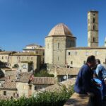 Anziani seduti su panchine nella piazza di un borgo toscano, intenti a raccontarsi storie.